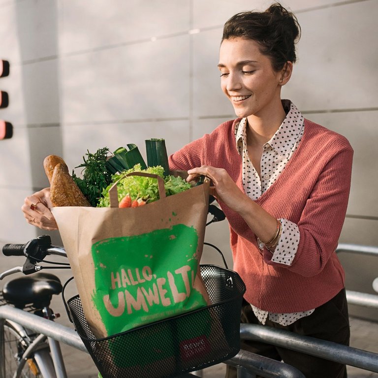 Eine lächelnde Frau platziert ihre REWE Einkaufstüte in ihrem Fahrradkorb. Das Setting ist draußen an festen Fahrradständern vor einem REWE Gebäude. 